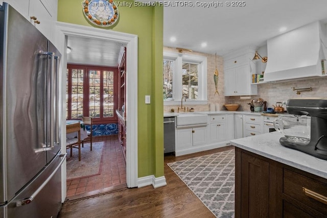 kitchen with sink, white cabinetry, stainless steel appliances, custom range hood, and decorative backsplash