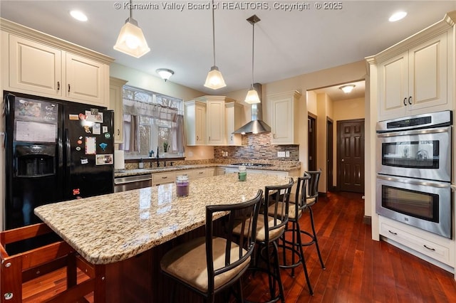 kitchen featuring wall chimney range hood, a kitchen bar, stainless steel appliances, a kitchen island, and decorative light fixtures