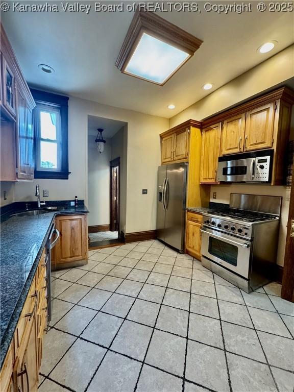 kitchen with light tile patterned flooring, a sink, appliances with stainless steel finishes, brown cabinetry, and dark countertops