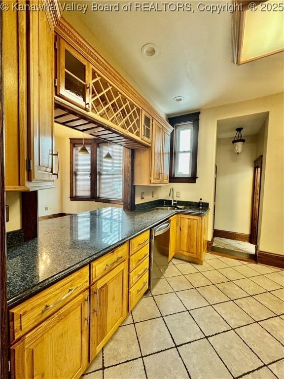 kitchen with light tile patterned flooring, baseboards, stainless steel dishwasher, brown cabinetry, and glass insert cabinets