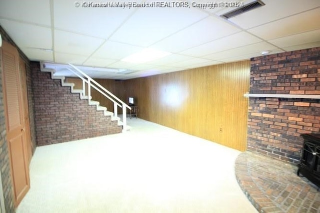 finished basement with visible vents, brick wall, stairway, a wood stove, and a paneled ceiling