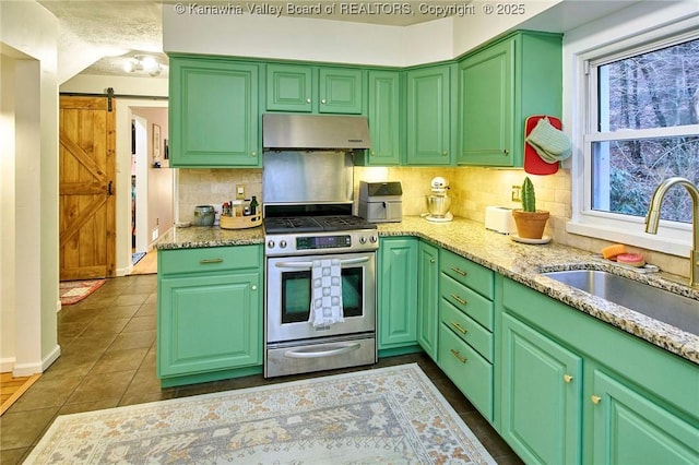 kitchen with a barn door, stainless steel gas stove, a sink, under cabinet range hood, and dark tile patterned floors