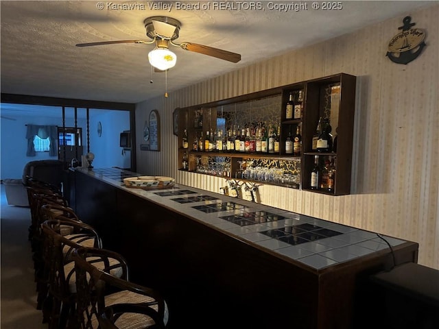 bar featuring wallpapered walls, ceiling fan, bar area, and a textured ceiling