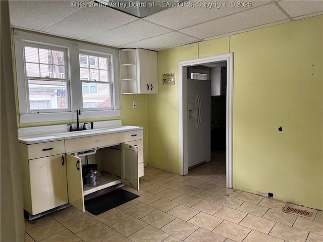 kitchen with white refrigerator with ice dispenser, open shelves, light countertops, a sink, and a drop ceiling