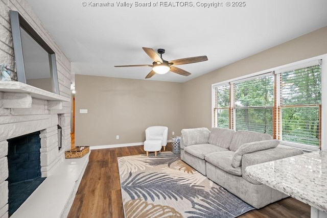 living area with ceiling fan, baseboards, a stone fireplace, and wood finished floors