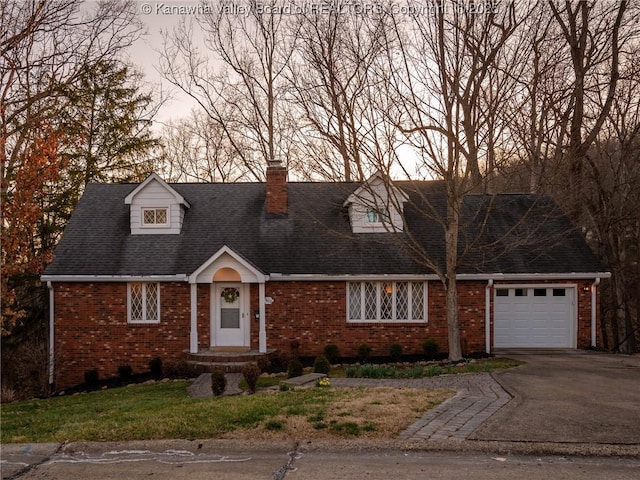 new england style home with driveway, roof with shingles, a garage, brick siding, and a chimney