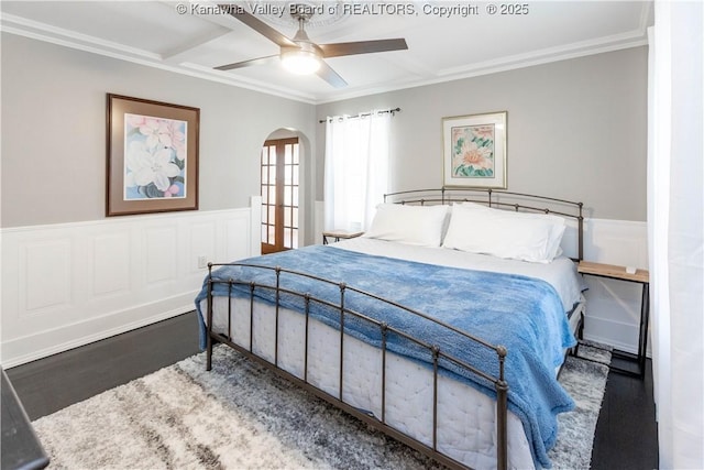 bedroom featuring a ceiling fan, wood finished floors, arched walkways, ornamental molding, and wainscoting