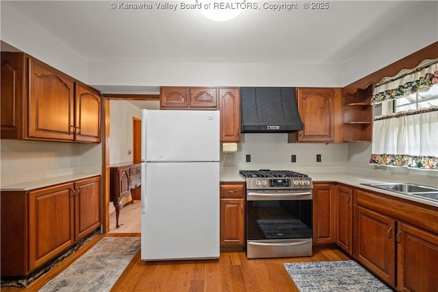 kitchen featuring wall chimney range hood, light countertops, freestanding refrigerator, light wood-style floors, and stainless steel gas range