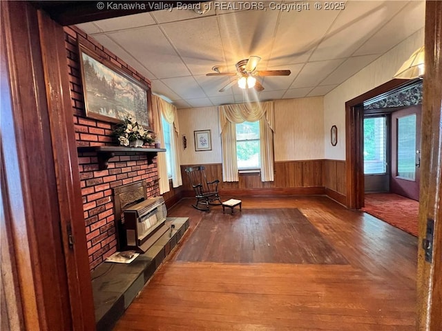 unfurnished living room with a ceiling fan, wood finished floors, wooden walls, wainscoting, and a brick fireplace