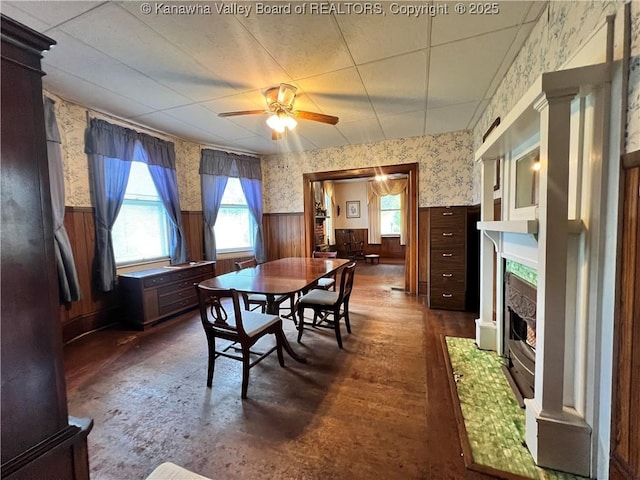 dining space featuring wallpapered walls, ceiling fan, a fireplace with flush hearth, and wainscoting