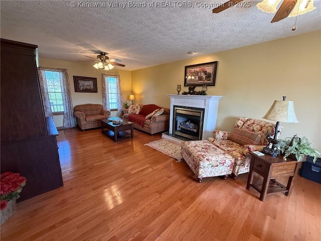living room featuring a fireplace with raised hearth, a textured ceiling, ceiling fan, and light wood-style flooring
