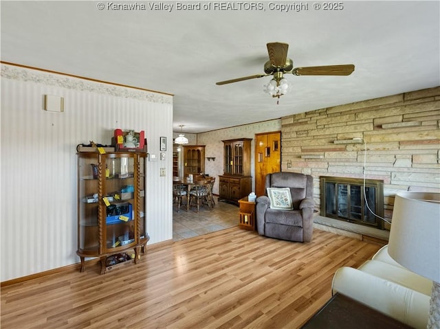 living room featuring a stone fireplace, a ceiling fan, wallpapered walls, and wood finished floors