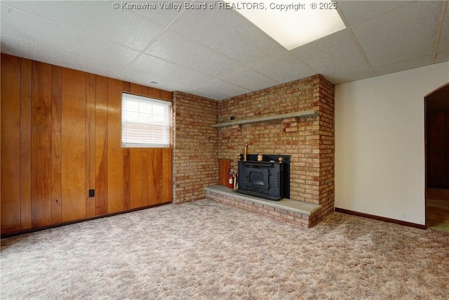 unfurnished living room featuring a wood stove, a drop ceiling, carpet, wood walls, and baseboards