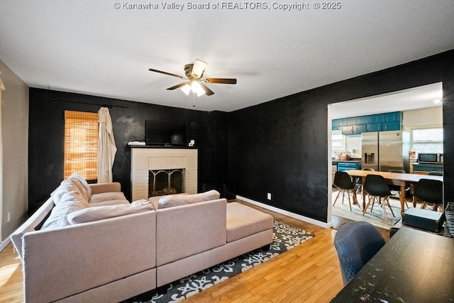 living room with a ceiling fan, light wood finished floors, a brick fireplace, and baseboards