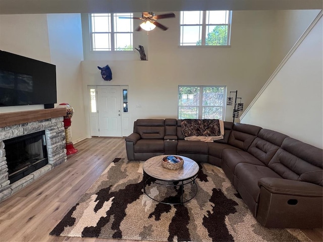 living room featuring ceiling fan, a stone fireplace, light wood-type flooring, and a high ceiling