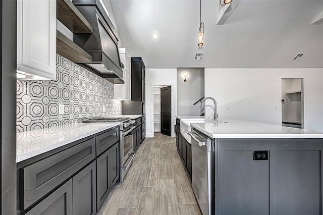 kitchen featuring appliances with stainless steel finishes, backsplash, a kitchen island with sink, wall chimney range hood, and pendant lighting