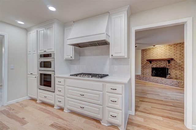 kitchen with appliances with stainless steel finishes, premium range hood, a brick fireplace, white cabinets, and light hardwood / wood-style floors