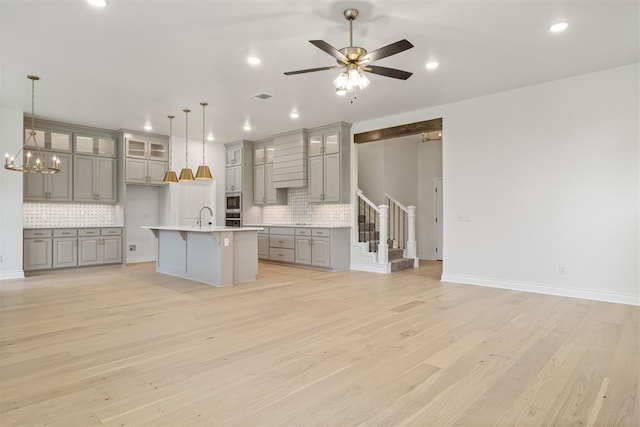 kitchen featuring ceiling fan with notable chandelier, decorative backsplash, light wood-type flooring, an island with sink, and decorative light fixtures