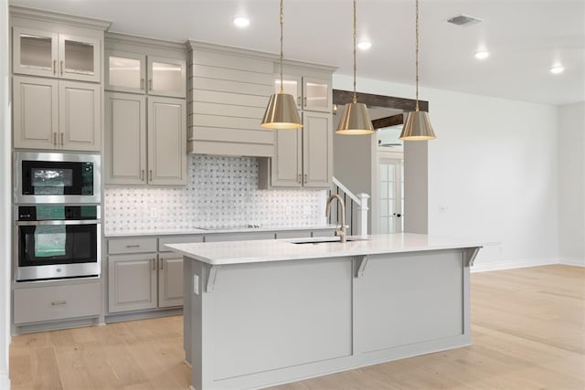 kitchen with sink, light hardwood / wood-style flooring, decorative light fixtures, a kitchen island with sink, and appliances with stainless steel finishes