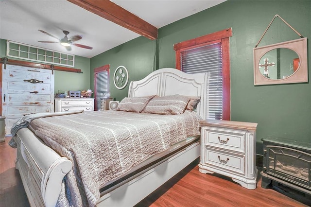 bedroom with beamed ceiling, wood-type flooring, a wood stove, and ceiling fan