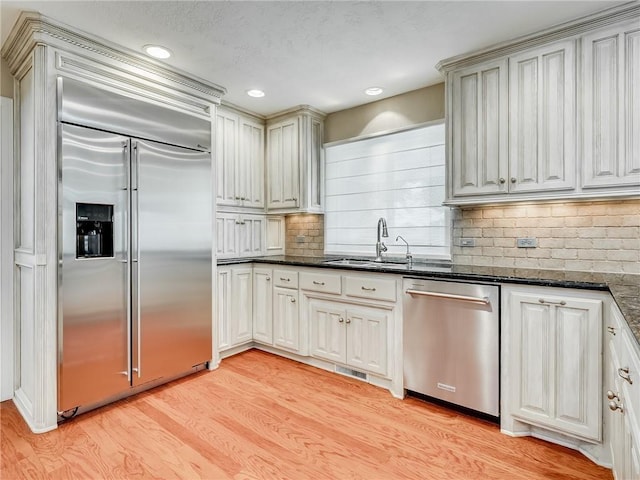 kitchen featuring sink, stainless steel appliances, backsplash, dark stone countertops, and light wood-type flooring