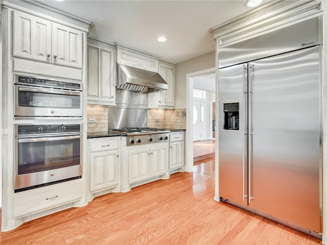 kitchen with decorative backsplash, light wood-type flooring, stainless steel appliances, wall chimney range hood, and dark stone countertops