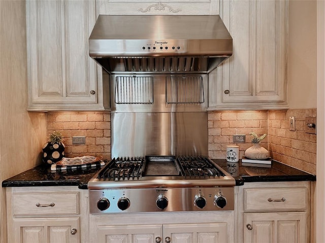 kitchen featuring wall chimney exhaust hood, dark stone countertops, cream cabinets, stainless steel gas stovetop, and decorative backsplash