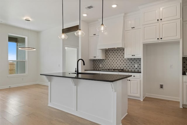 kitchen with custom exhaust hood, a kitchen island with sink, sink, light wood-type flooring, and white cabinetry