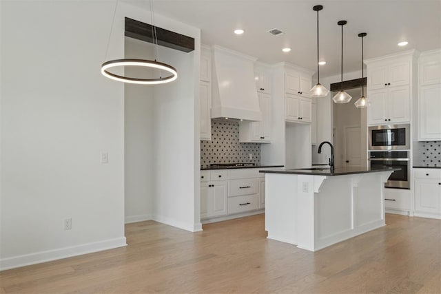 kitchen with backsplash, premium range hood, an island with sink, light hardwood / wood-style floors, and stainless steel appliances