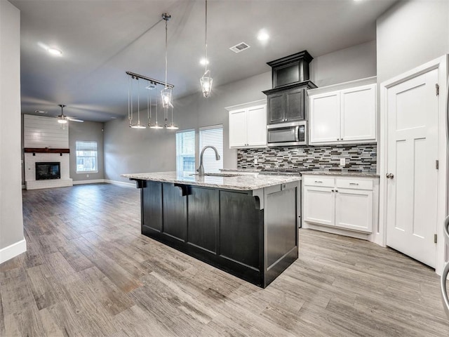 kitchen with a kitchen island with sink, ceiling fan, a fireplace, decorative light fixtures, and white cabinetry