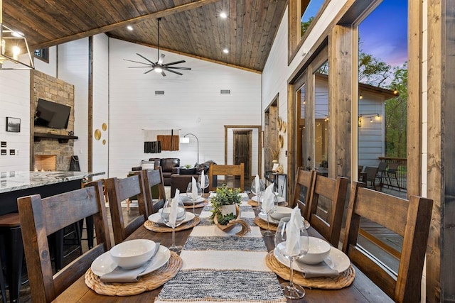 dining room with an outdoor stone fireplace, ceiling fan, wood ceiling, and high vaulted ceiling