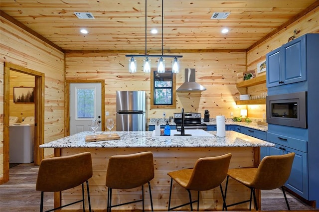 kitchen with wooden ceiling, wall chimney range hood, blue cabinetry, an island with sink, and stainless steel appliances