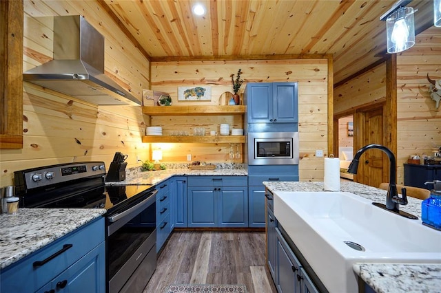 kitchen featuring wall chimney exhaust hood, stainless steel appliances, blue cabinets, sink, and wood walls