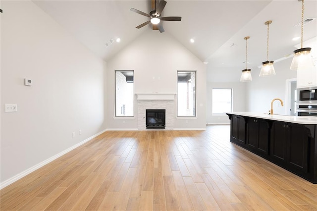 unfurnished living room featuring a stone fireplace, high vaulted ceiling, sink, ceiling fan, and light hardwood / wood-style floors