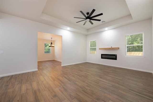 unfurnished living room featuring plenty of natural light, dark hardwood / wood-style floors, ceiling fan with notable chandelier, and a tray ceiling