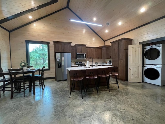 kitchen featuring appliances with stainless steel finishes, stacked washing maching and dryer, beam ceiling, high vaulted ceiling, and an island with sink