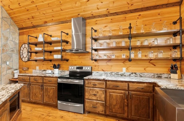 kitchen featuring wall chimney range hood, light stone counters, light hardwood / wood-style flooring, stainless steel electric range, and wooden walls