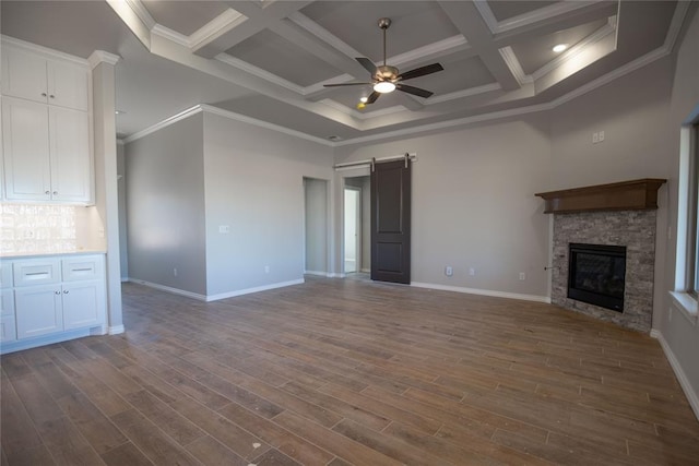 unfurnished living room featuring coffered ceiling, a barn door, a stone fireplace, dark hardwood / wood-style flooring, and crown molding