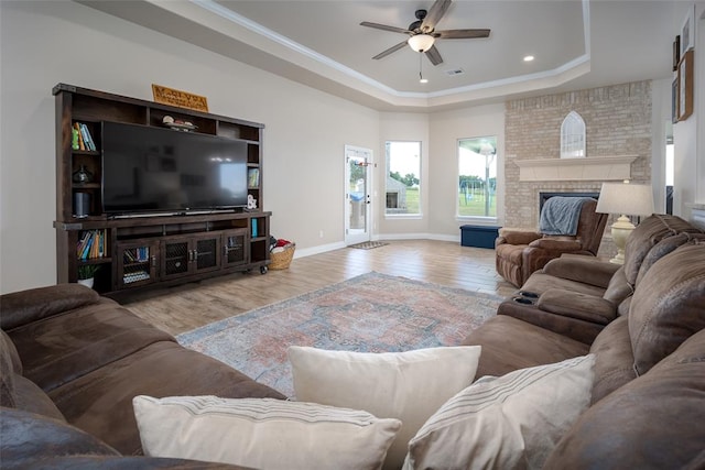 living room featuring light wood-type flooring, a brick fireplace, a tray ceiling, ceiling fan, and crown molding