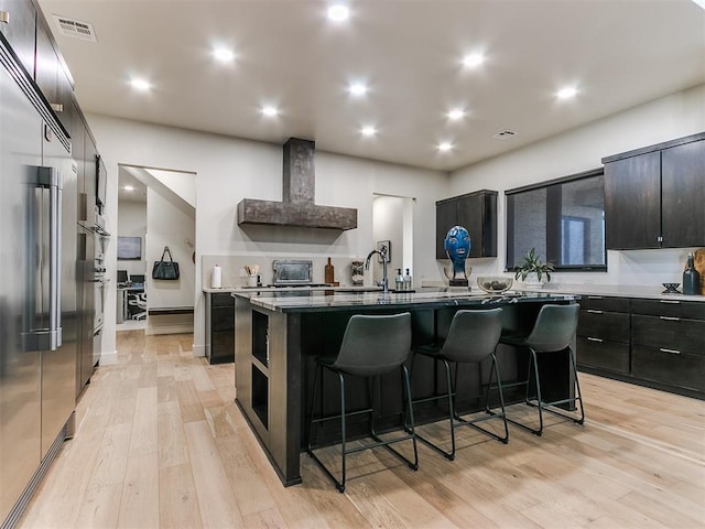 kitchen with wall chimney range hood, light hardwood / wood-style flooring, an island with sink, stainless steel built in refrigerator, and a breakfast bar area