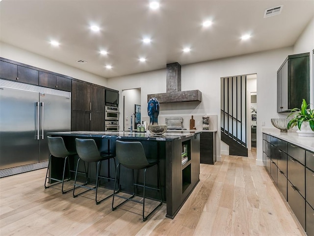 kitchen featuring wall chimney range hood, a large island with sink, a breakfast bar area, appliances with stainless steel finishes, and light wood-type flooring