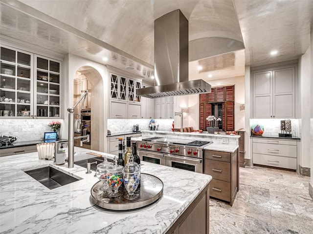 kitchen featuring backsplash, island exhaust hood, a center island with sink, and range with two ovens