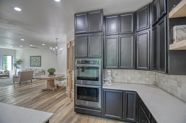 kitchen featuring light stone counters, light wood-type flooring, a notable chandelier, stainless steel double oven, and decorative backsplash