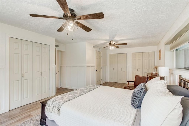 bedroom featuring a textured ceiling, ceiling fan, a wainscoted wall, multiple closets, and light wood-type flooring