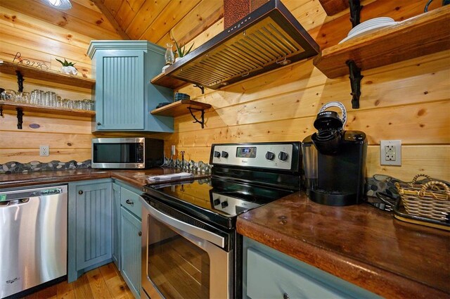 kitchen featuring appliances with stainless steel finishes, blue cabinets, exhaust hood, lofted ceiling, and wood walls