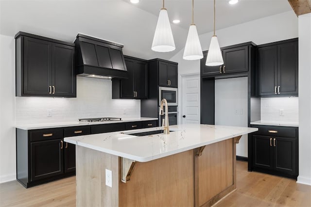 kitchen featuring appliances with stainless steel finishes, custom range hood, decorative light fixtures, light hardwood / wood-style floors, and an island with sink