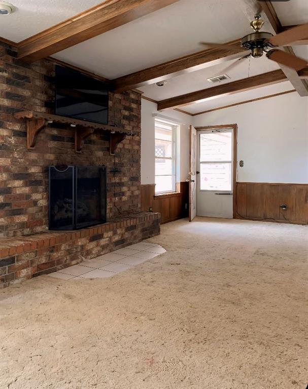 unfurnished living room featuring wood walls, a brick fireplace, ceiling fan, beamed ceiling, and carpet floors