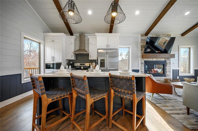 kitchen featuring appliances with stainless steel finishes, plenty of natural light, a center island with sink, vaulted ceiling with beams, and wall chimney exhaust hood