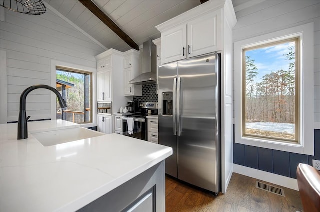 kitchen with white cabinetry, sink, lofted ceiling with beams, appliances with stainless steel finishes, and wall chimney exhaust hood