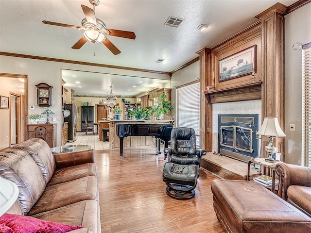 living room with a tiled fireplace, ceiling fan, crown molding, and light wood-type flooring